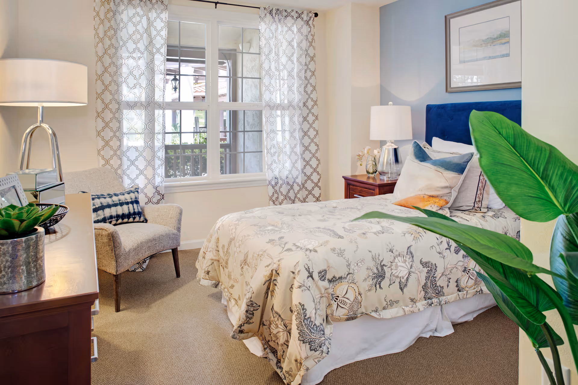 Bright, well-decorated bedroom with a bed, bedside tables and lamps, an armchair by a window with patterned curtains, and a large potted plant.