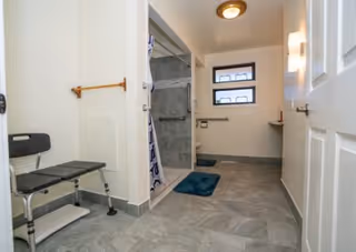 A spacious bathroom with a walk-in shower featuring gray tiles and a shower curtain. There is a black shower chair with metal legs, a towel rack on the wall, two small windows, a blue bath mat on the floor, and a white door partially open.