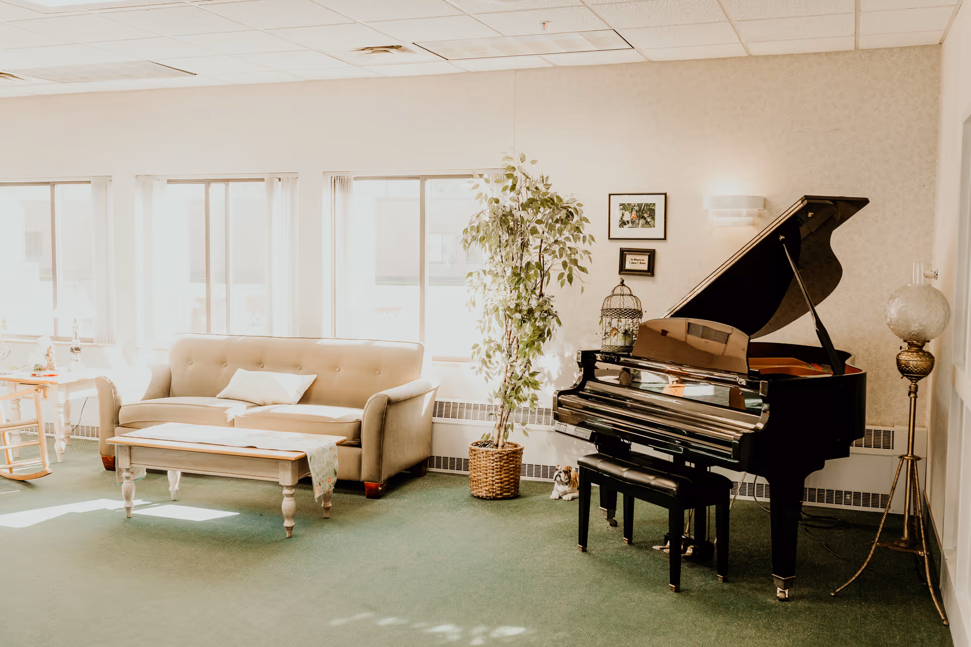A bright and cozy living room area with a beige sofa, a wooden coffee table, a potted plant, and a black grand piano with a matching bench. The room has large windows letting in natural light and light-colored walls with framed pictures. There is also a vintage floor lamp with a glass globe shade.