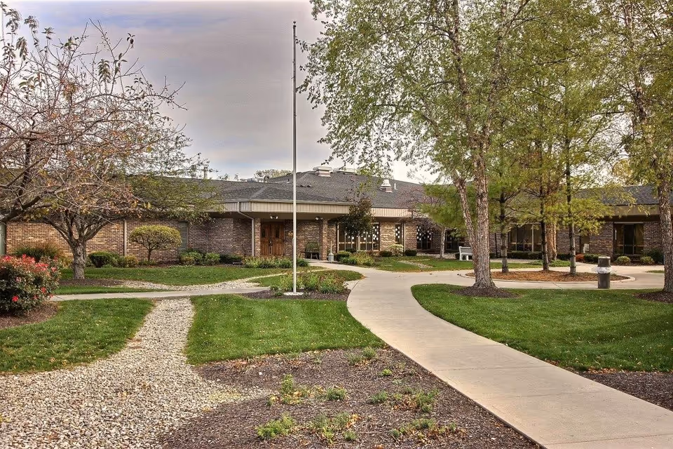 Outdoor view of Greenwood Healthcare Center showing a single-story brick building surrounded by green lawns, trees, and landscaped pathways under a cloudy sky.