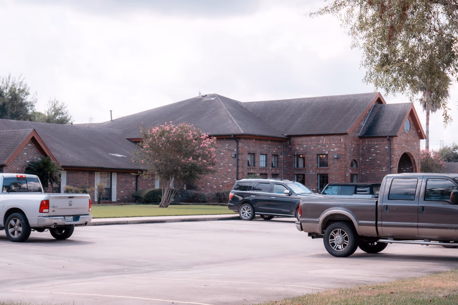 Parking lot with several parked vehicles in front of a large brick building with a sloped roof and multiple windows. There are trees and shrubs around the building, and the sky is cloudy.