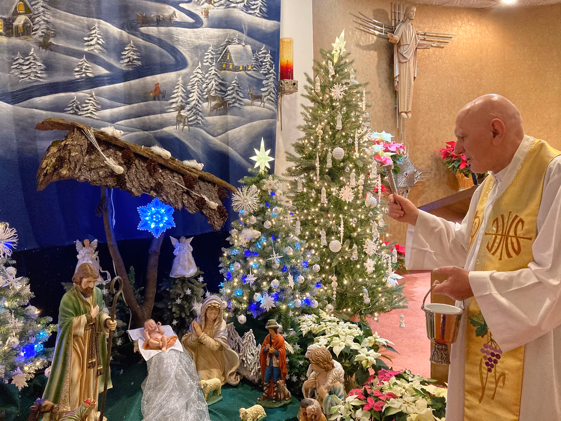 A priest in white and gold vestments is blessing a nativity scene with a holy water sprinkler inside a room decorated for Christmas. The nativity scene includes figurines of Mary, Joseph, baby Jesus, and other characters, surrounded by decorated Christmas trees with white and blue lights and ornaments. The background features a winter-themed mural and poinsettia plants.