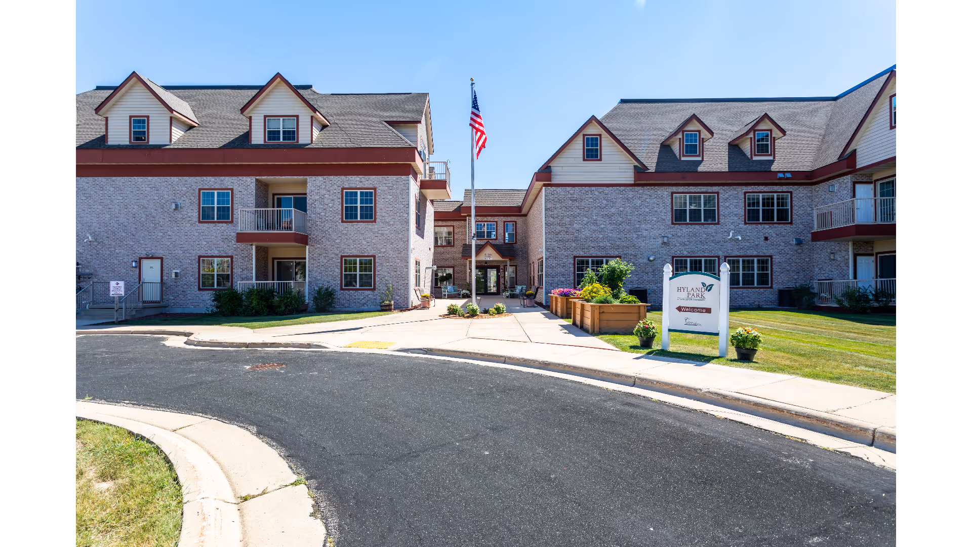 Front entrance of a two-story brick senior living building with balconies, landscaping, a sidewalk sign, and an American flag.