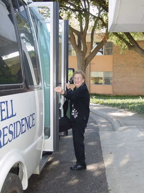 An elderly woman smiling as she steps onto a white shuttle bus with the words 'Stilwell Residence' partially visible on the side. The scene is outdoors with a building, trees, and greenery in the background.