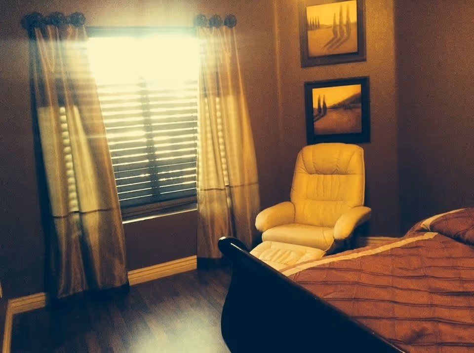 A cozy bedroom with a large window covered by curtains and blinds, a comfortable beige armchair, two framed landscape paintings on the wall, and a bed with a brown quilted bedspread.