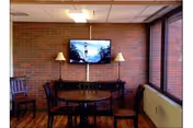 A small dining area with a round wooden table and four chairs, two floor lamps on either side of a wall-mounted flat screen TV, and large windows on the right side letting in natural light. The walls are brick and the floor is wooden.