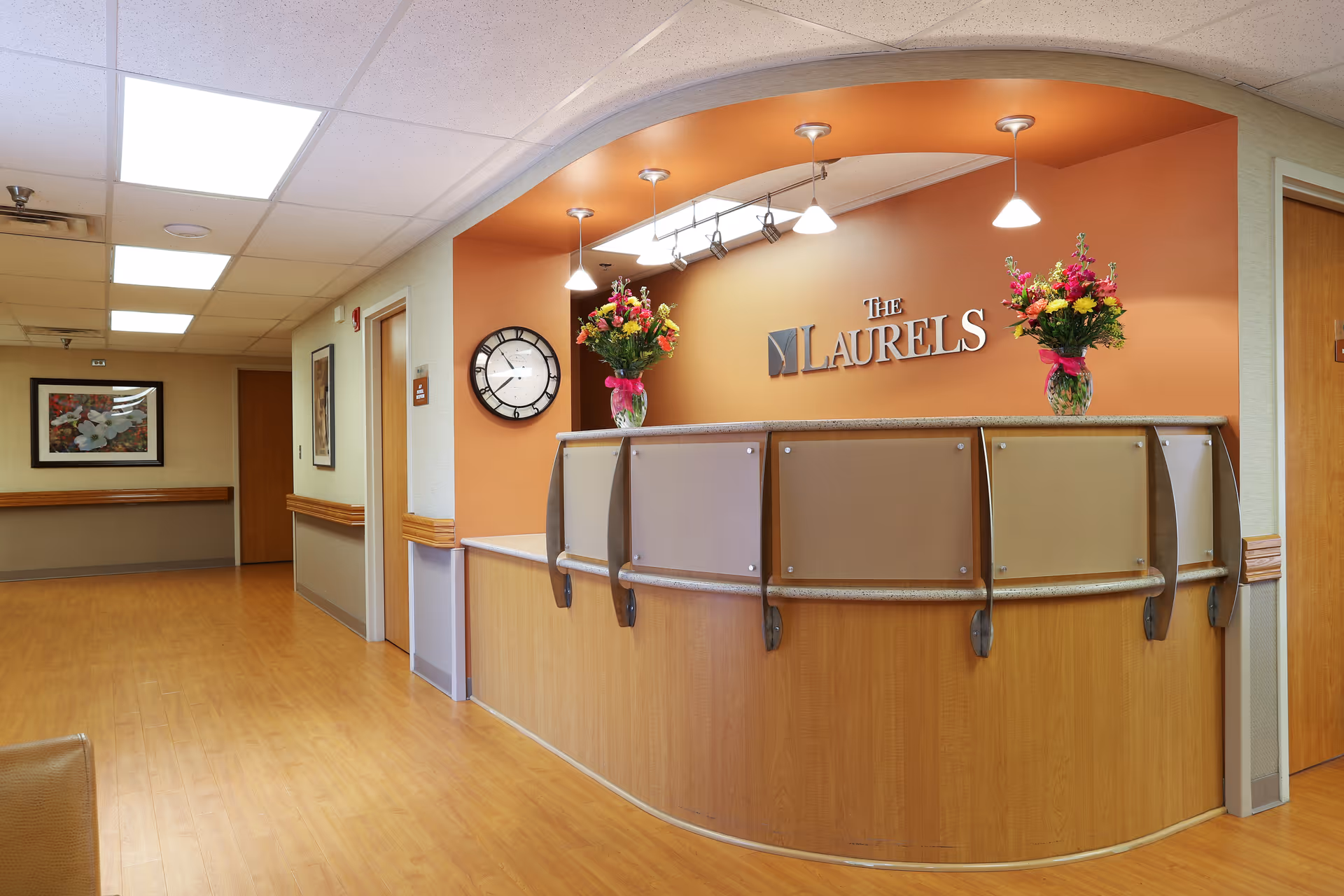 Reception area of The Laurels senior living facility with a curved wooden front desk, two vases of colorful flowers on the counter, a wall clock, and a sign that reads 'The Laurels' on an orange accent wall. The hallway adjacent to the reception has framed artwork and wooden doors.