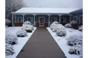 Snow-covered walkway leading to the front entrance of a single-story building with a covered porch and shrubs.