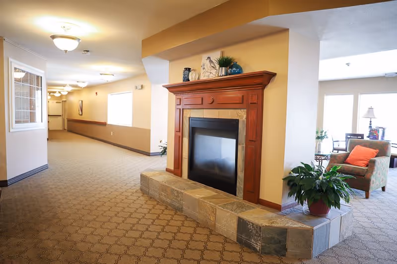 A well-lit hallway in a senior living facility with beige walls and patterned carpet. There is a decorative fireplace with a wooden mantle and stone base on the right side, adorned with small plants and decorative items. Beyond the fireplace, a cozy seating area with an armchair and a side table with a lamp is visible near large windows letting in natural light.