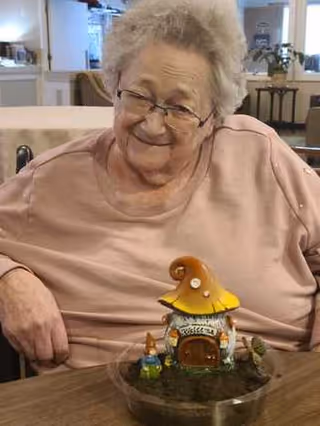 An elderly woman with glasses and short gray hair is sitting at a table indoors, smiling. In front of her on the table is a small decorative mushroom house with tiny figurines around it.