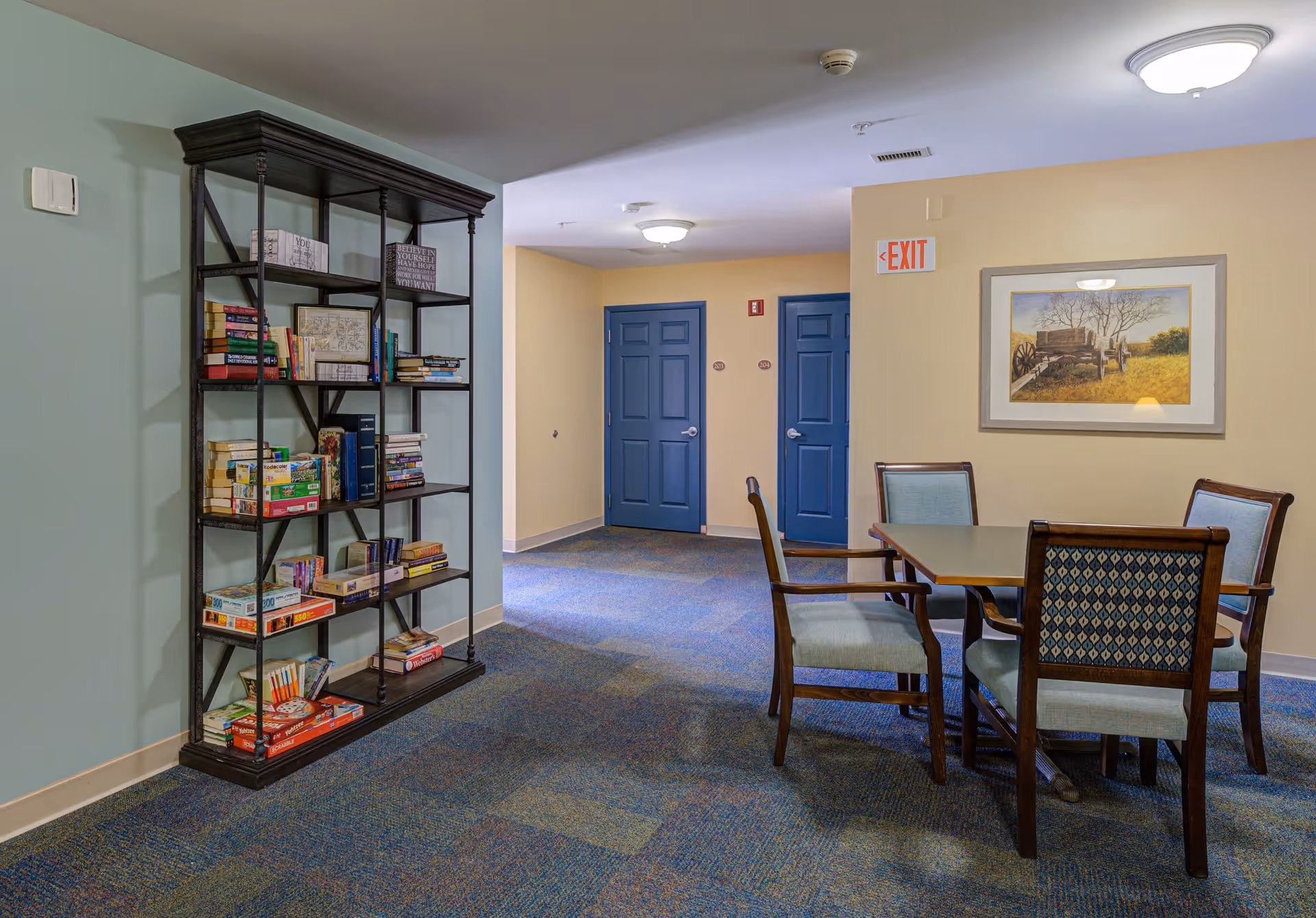 Interior view of a senior living community common area featuring a tall black bookshelf filled with books and board games on the left, a table with four chairs on the right, blue doors in the background, a framed painting on the wall, and an exit sign above the doors.