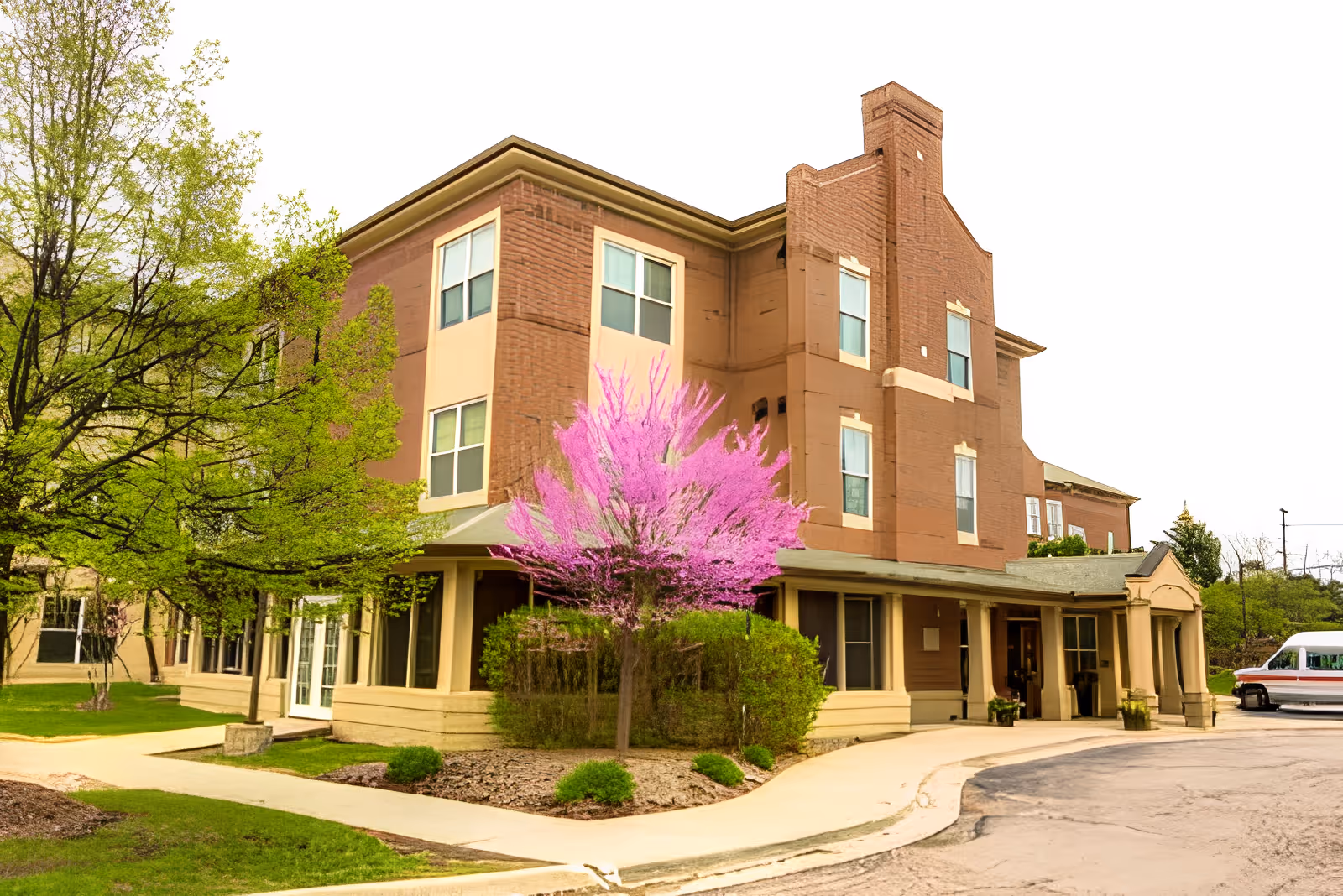 Brick three-story senior living building with a covered entrance, landscaped grounds, and a blooming pink tree in front.