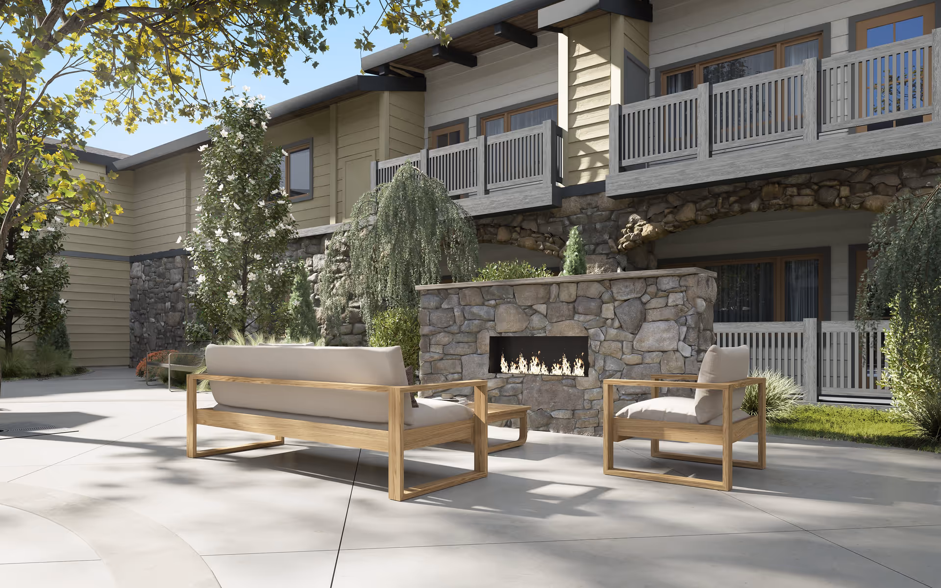 Outdoor patio area at Hearthstone Lodge Senior Residences featuring cushioned wooden seating arranged around a stone fireplace. The building exterior has stone and wood siding with balconies and windows. Trees and shrubs provide greenery around the patio.