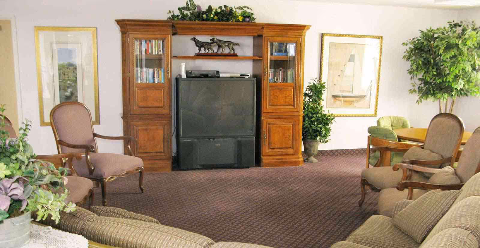 A senior living facility common area with a large wooden entertainment center holding a television and books. The room has multiple upholstered chairs and sofas arranged around the space, with framed artwork on the walls and potted plants adding greenery.