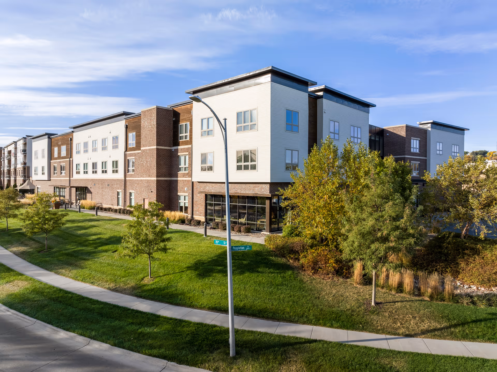 Exterior view of a modern multi-story senior living facility building with brick and light-colored siding, surrounded by green lawns, trees, and a sidewalk under a partly cloudy sky.