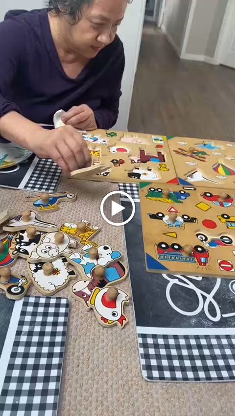 A person is sitting at a table playing with several wooden peg puzzles featuring colorful animal and vehicle shapes. The table has a checkered and solid color tablecloth, and the background shows a hallway with wooden flooring.