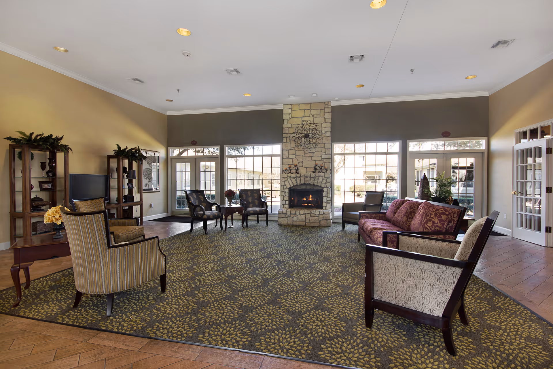 A spacious living room area with a patterned carpet, several upholstered chairs and a sofa arranged around a stone fireplace. Large windows and glass doors let in natural light, and there are wooden shelves with decorative items and a television on the left side.