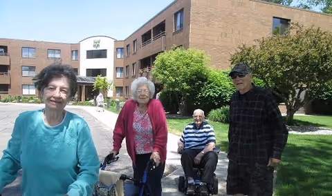 Four elderly individuals outside on a sunny day in front of a multi-story brick building with greenery and a paved walkway. One woman in a turquoise sweater is smiling at the camera, another woman in a pink cardigan is using a walker, a man in a striped shirt is seated in a mobility scooter, and another man wearing sunglasses and a dark plaid shirt stands nearby.