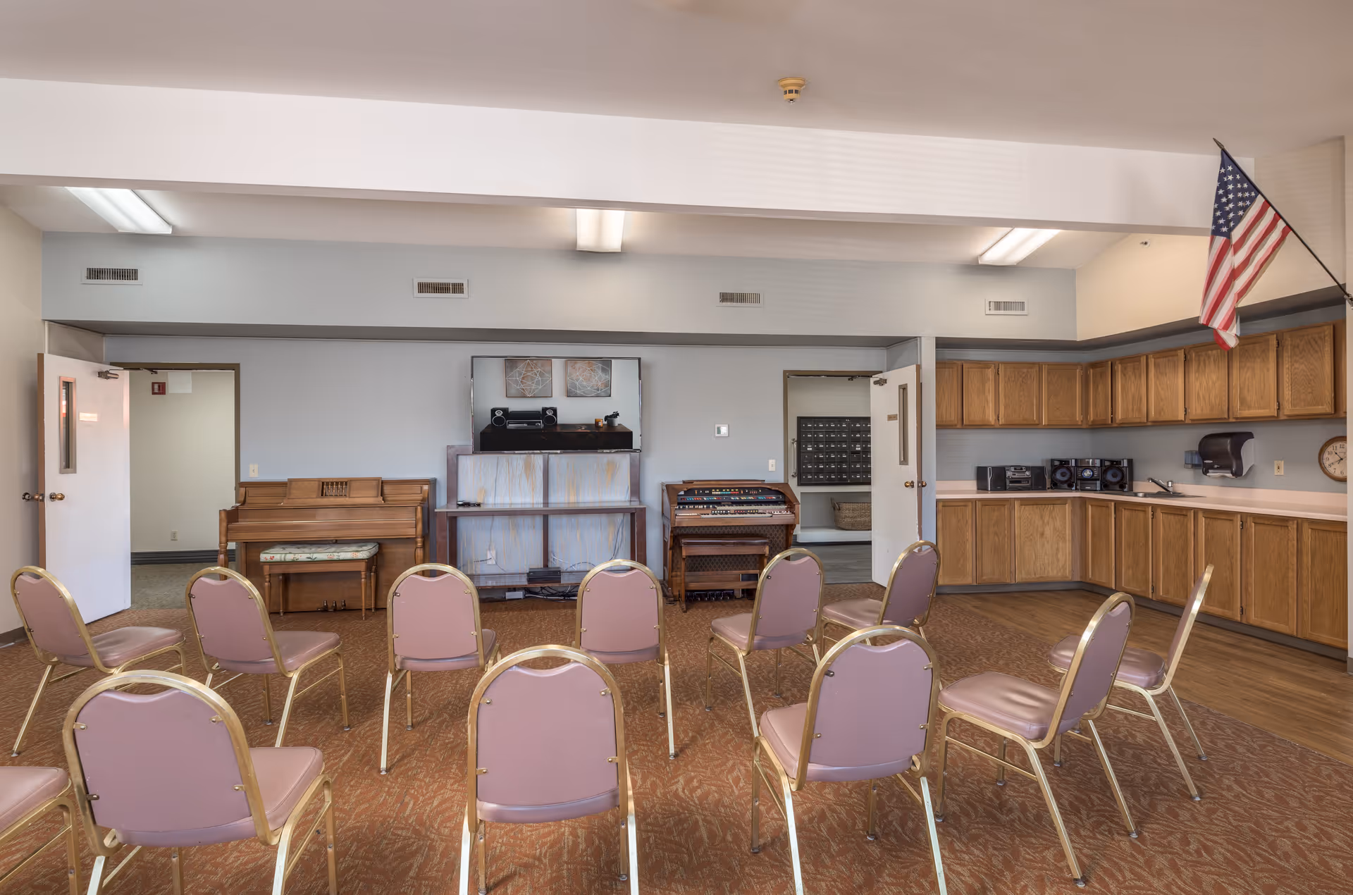 An interior activity room with arranged chairs facing a piano and organ, a kitchenette along one wall, and an American flag.