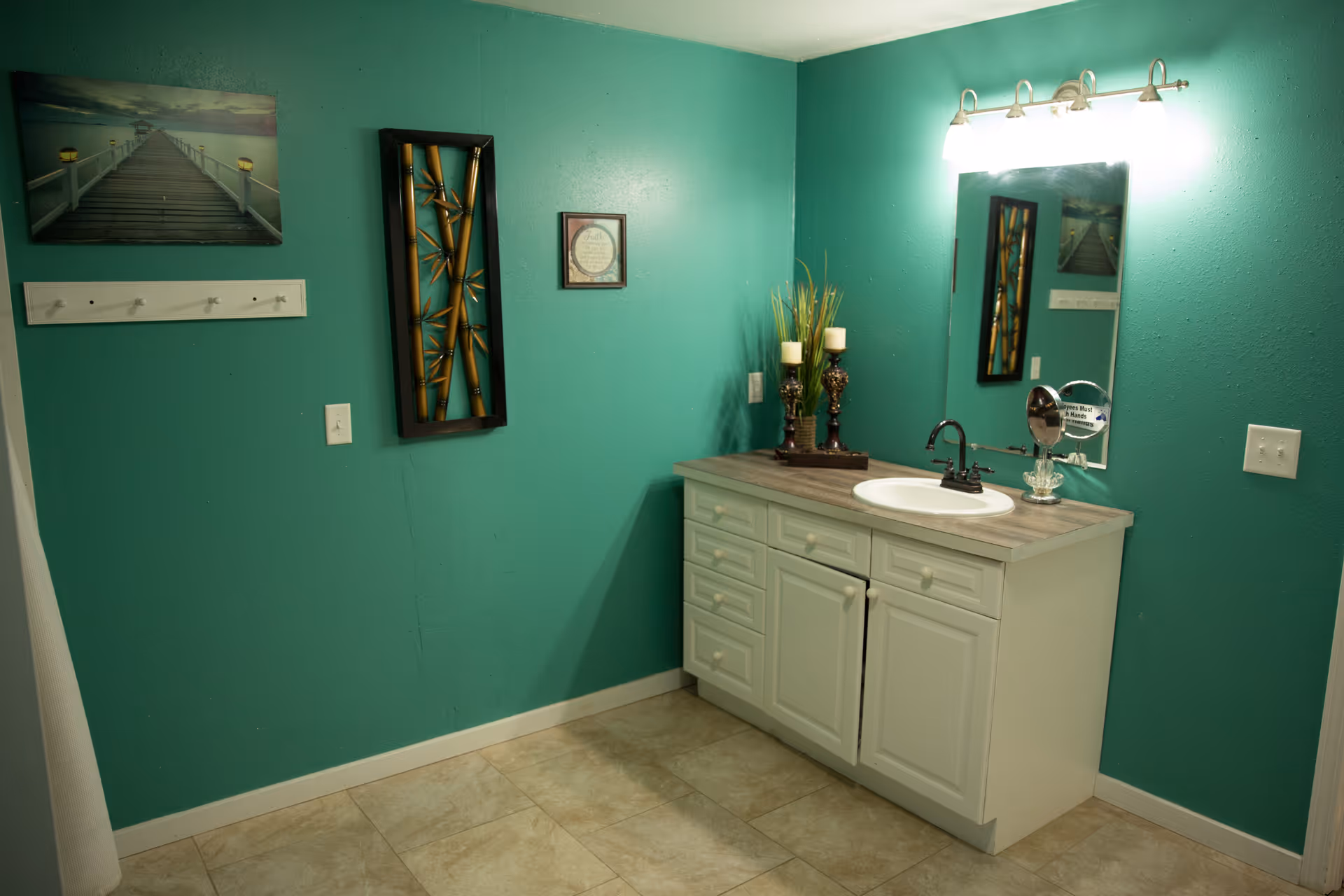 A bathroom with teal walls featuring a white vanity with a sink, a mirror above it, and a light fixture. The vanity countertop has decorative candles and a small mirror. The wall has framed artwork and a coat rack with hooks. The floor is tiled in a beige color.