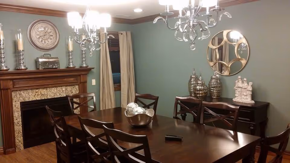 Dining room with a large wooden table and chairs beneath chandeliers, a fireplace mantel, and decorative vases and a mirror on green walls.