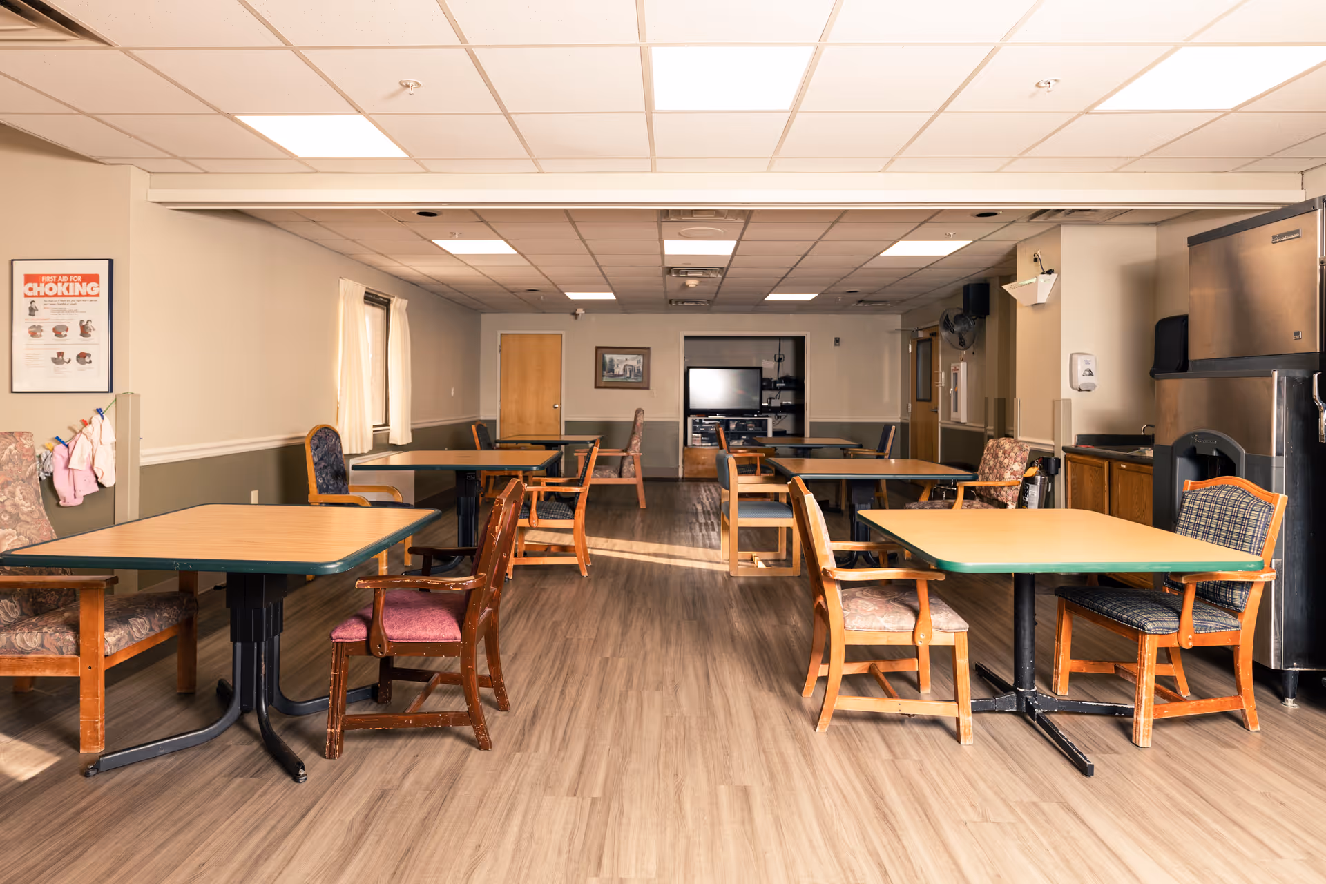 A dining room with several wooden tables and chairs arranged neatly. The room has a light-colored ceiling with recessed lighting, light beige walls with a darker green wainscoting, and wood-patterned flooring. On the left wall, there is a framed poster about first aid for choking. In the background, there is a TV and some cabinets. A large stainless steel refrigerator is visible on the right side of the image.