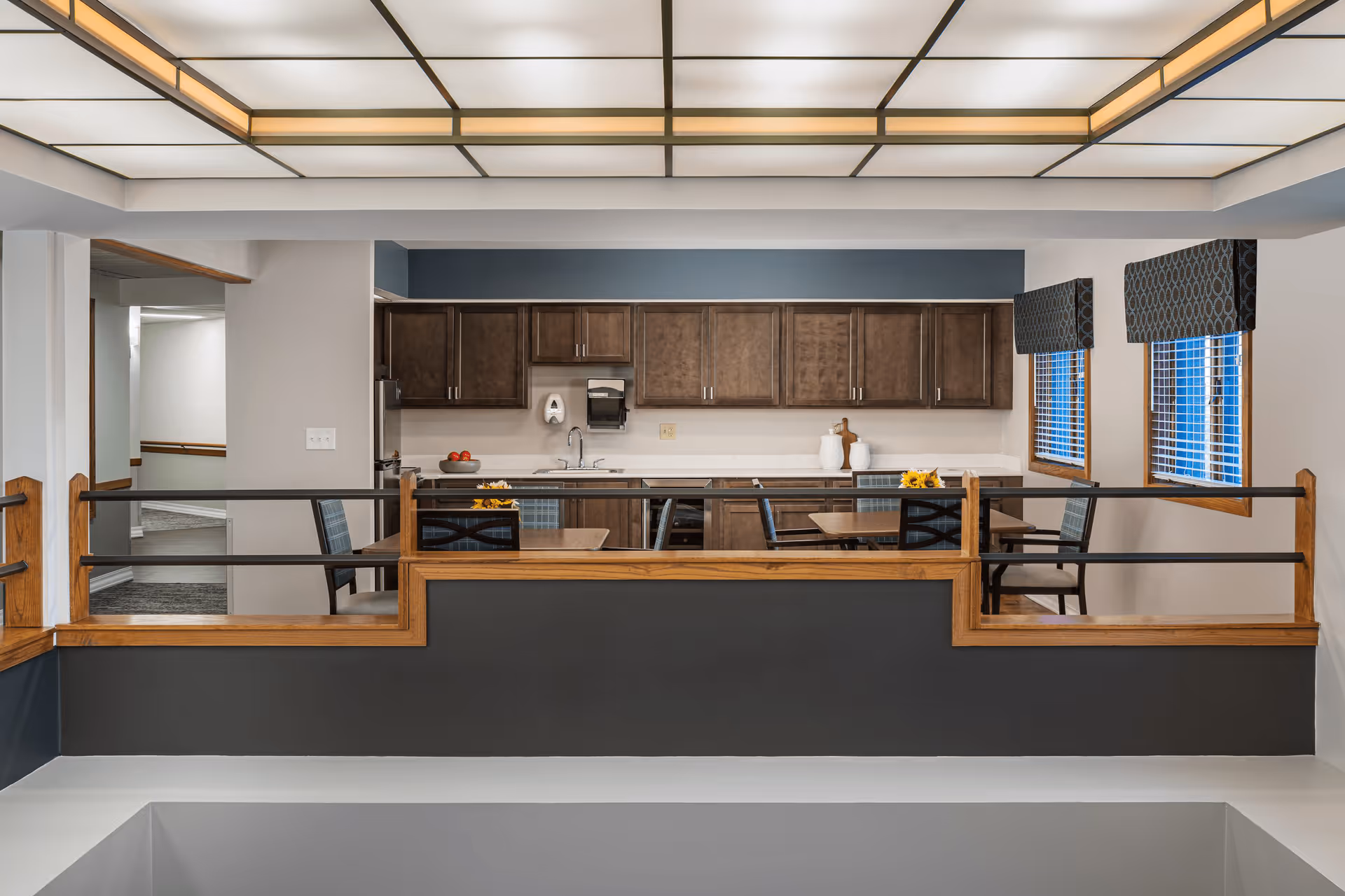 Interior view of a senior living community kitchen and dining area with wooden cabinets, a sink, and tables with chairs. The room has a modern ceiling light fixture and two windows with patterned valances.