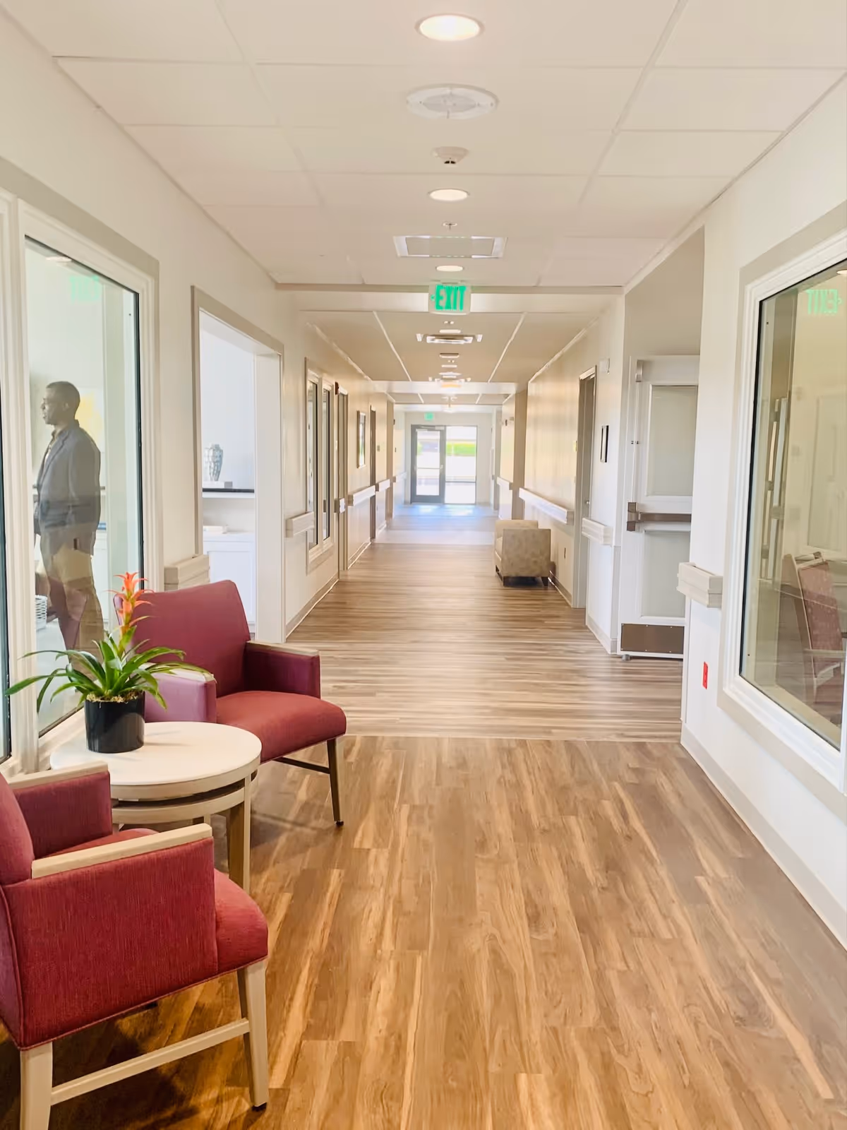 A bright, clean hallway in a senior living facility with wood-patterned flooring and white walls. Two red upholstered chairs and a small round table with a potted plant are positioned near a large window on the left. The hallway extends into the distance with doors and handrails along the walls, and an exit door visible at the far end. A person is standing outside the window on the left side.