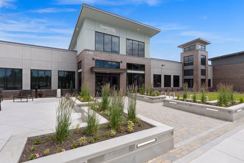 Front exterior of a modern senior living facility with a paved courtyard, raised planters, and seating.