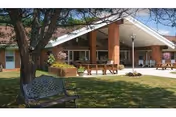 Outdoor view of Sena Kean Nursing and Rehabilitation Center showing a single-story brick building with a covered entrance. There are benches and tables on a paved area near the entrance, and a tree with a metal bench in the grassy foreground.