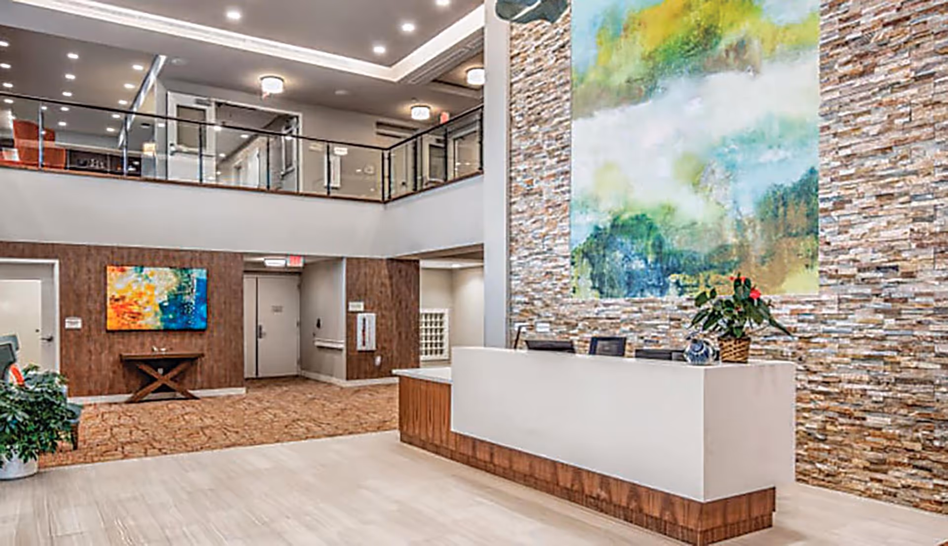 A spacious and modern reception area with a white and wood front desk, a large colorful abstract painting on a stone accent wall behind the desk, a potted plant on the desk, and a second floor balcony with glass railing overlooking the area. The floor is light-colored tile, and there is a carpeted area with a small table and another colorful painting on the far wall.