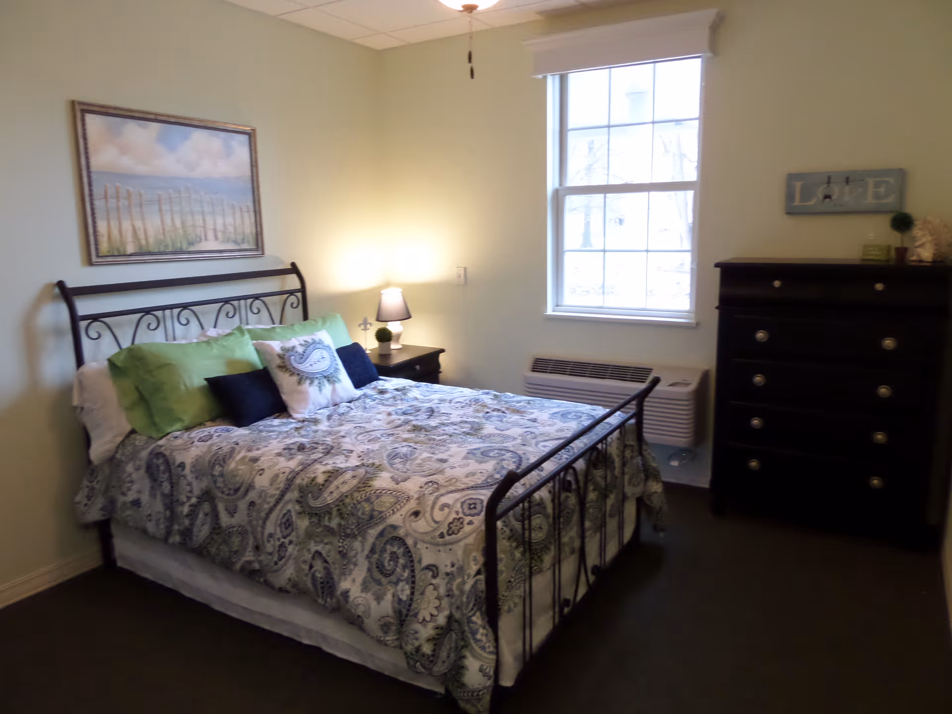 A bedroom with a metal frame bed covered with a paisley patterned comforter and multiple pillows. There is a nightstand with a lamp and a small plant next to the bed. A window with white trim lets in natural light. On the right side of the room is a dark wooden dresser with decorative items on top. A framed painting of a beach scene hangs on the wall above the bed.