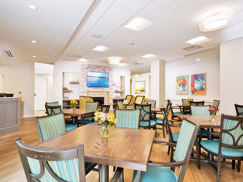 A bright and spacious dining room with multiple wooden tables and chairs featuring teal and striped upholstery. Each table has a small vase with fresh flowers. The room has light-colored walls adorned with colorful paintings and a mounted flat-screen TV above a fireplace. The ceiling has recessed lighting and modern circular light fixtures.