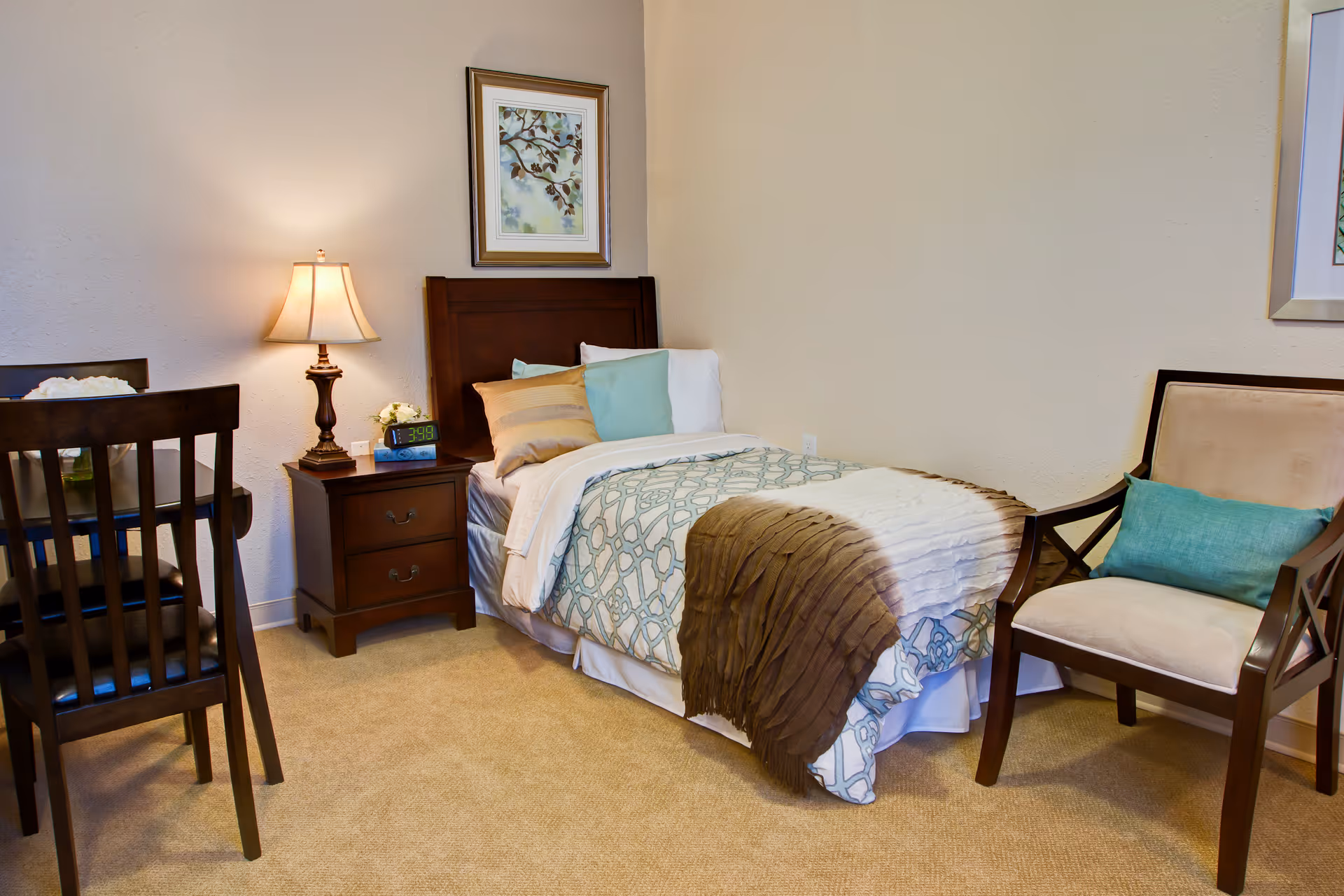 A cozy bedroom with a single bed featuring patterned bedding and multiple pillows. Next to the bed is a wooden nightstand with a lamp, a digital clock, and a small flower arrangement. To the right of the bed is a cushioned chair with a teal pillow. On the left side, part of a wooden dining table and chairs are visible. The walls are light-colored with framed artwork hanging above the bed and chair.