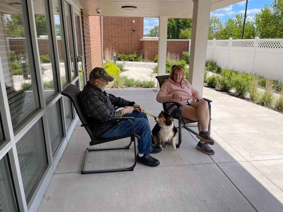 An elderly man and a woman sitting on chairs on a covered patio outside Prairie Pointe Assisted Living and Memory Care. The man is holding a leash attached to a small dog sitting between them. There is a garden with plants and a white fence in the background.