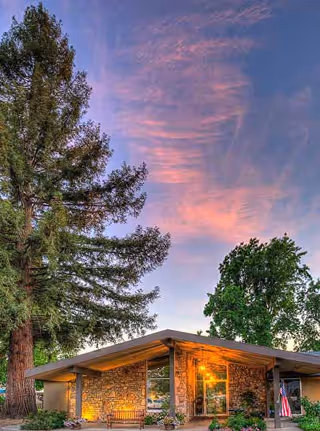 Stone-clad single-story entrance with warm exterior lights, a bench, American flag, and tall trees under a pink sunset sky.