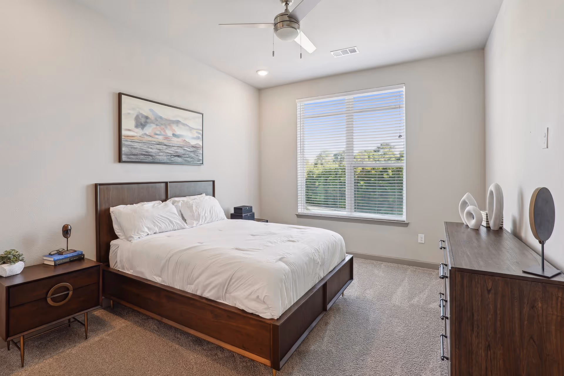 A bright bedroom with a large window covered by white blinds, a wooden bed frame with white bedding, a wooden nightstand with books and a small plant, a wooden dresser with decorative items, and a ceiling fan with light.