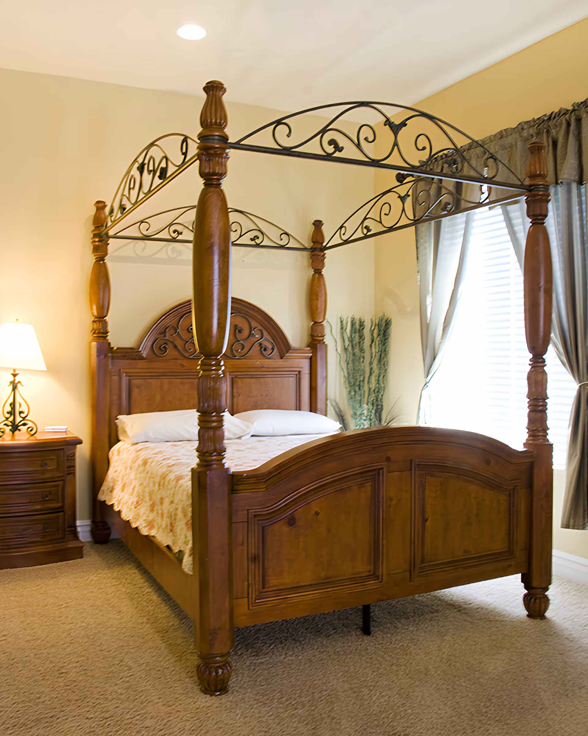 Sunlit bedroom with a wooden four-poster bed, nightstand with a lamp, and a curtained window.