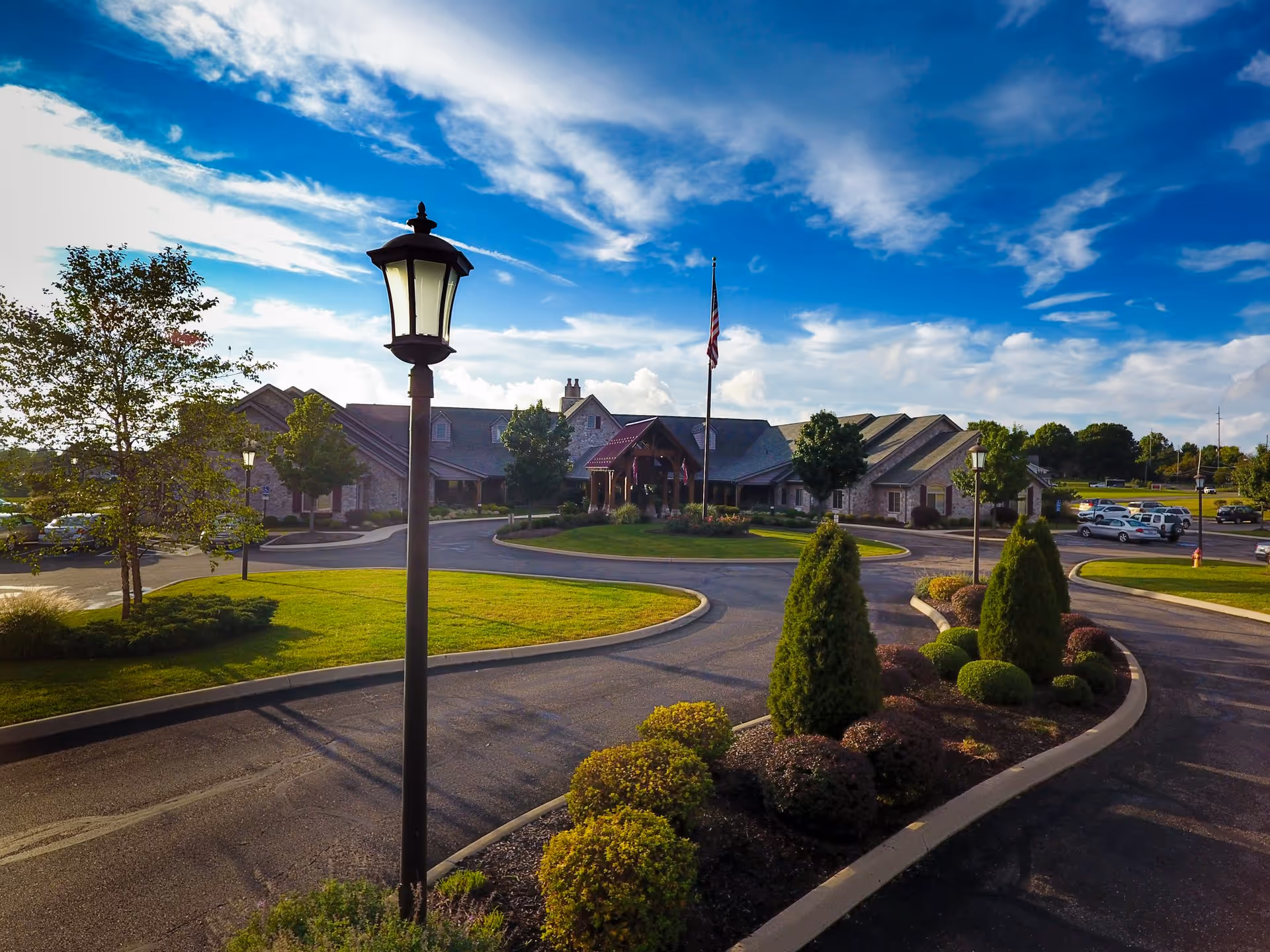 Exterior view of The Inn At University Village facility with a circular driveway, landscaped bushes and trees, a flagpole with an American flag, and several parked cars under a partly cloudy blue sky.