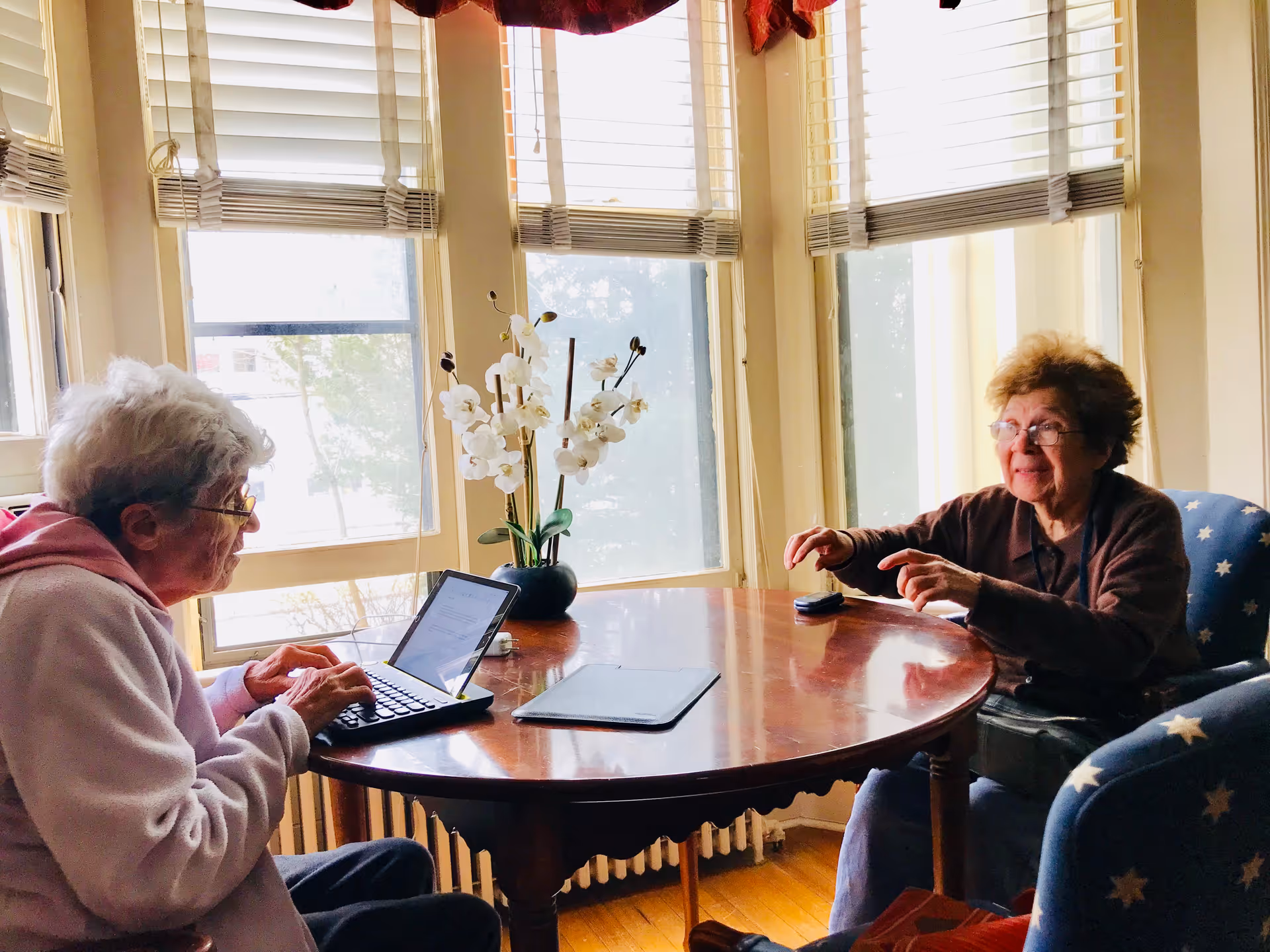 Two elderly women sit at a round wooden table by bay windows, one using a laptop while the other gestures.
