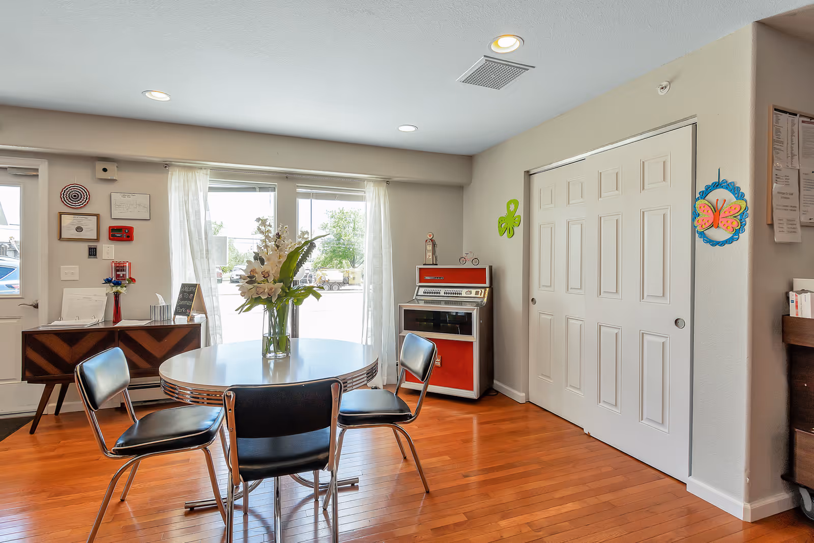 A bright room with wooden flooring featuring a round white table with a vase of flowers on top, surrounded by three black chairs with chrome legs. There is a large window with sheer white curtains letting in natural light. Against the wall is a small wooden cabinet with a retro red and white jukebox on top. The walls are decorated with colorful butterfly and flower wall hangings, and there are some framed certificates and notices near the door.