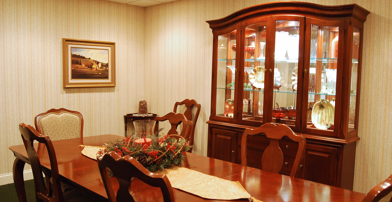 A traditional dining room with a polished wooden dining table and six matching chairs. The table is decorated with a festive centerpiece featuring red berries, pinecones, and greenery, along with a glass candle holder. Behind the table is a wooden china cabinet displaying various glassware and decorative plates. The walls have a light patterned wallpaper, and a framed landscape painting hangs on the wall.