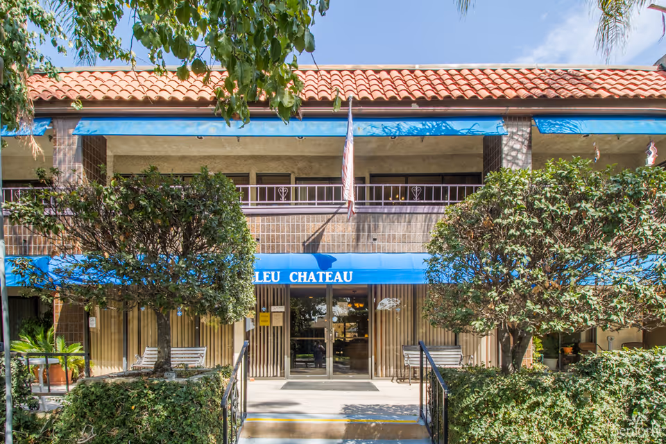 Front exterior view of a two-story building with a red tile roof and blue awnings. There are two trimmed trees and benches on either side of the entrance. An American flag is hanging above the entrance door.