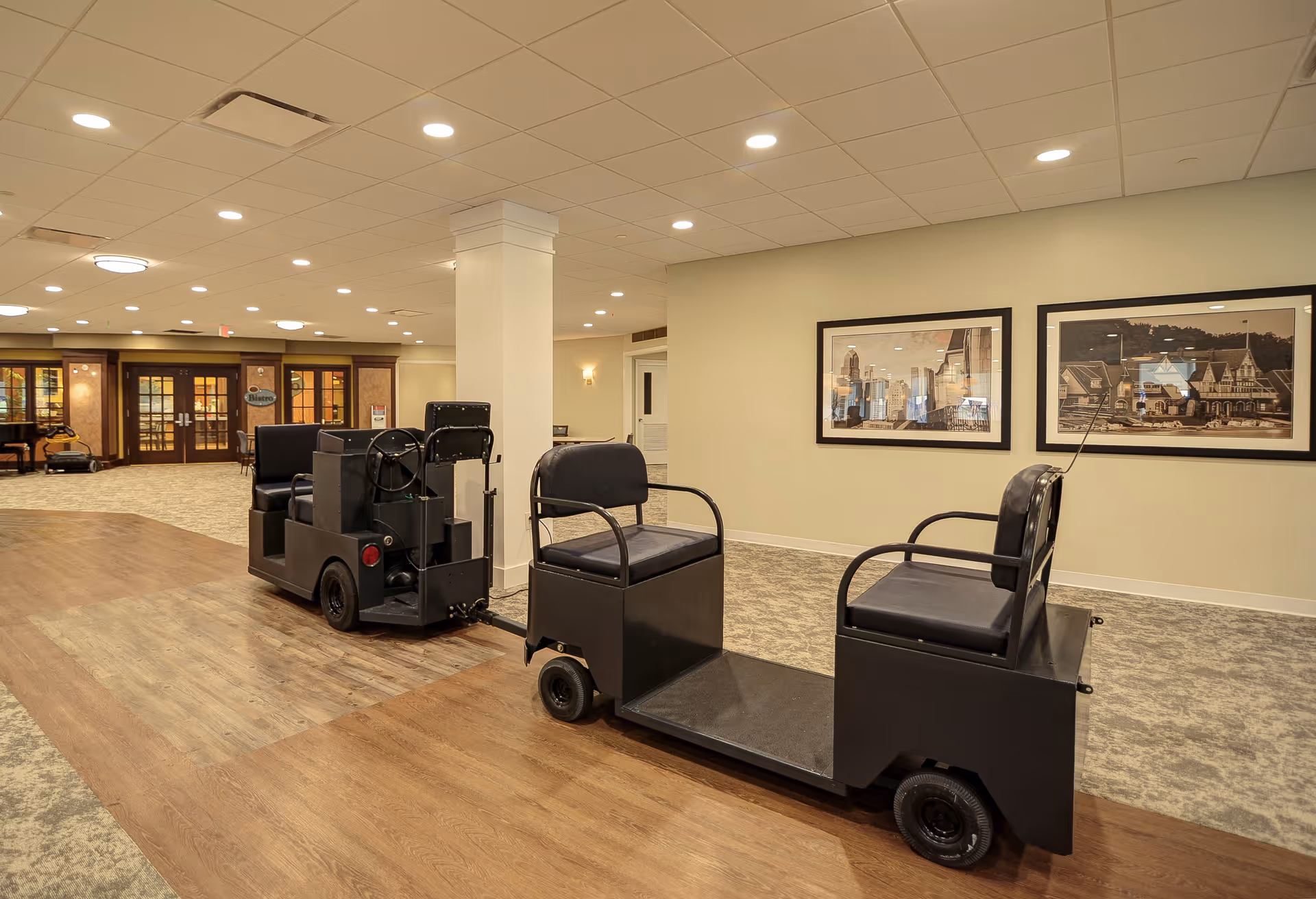 Interior view of a senior living facility hallway with a small electric cart designed to transport residents. The hallway has beige walls, carpeted floors with a wooden strip in the center, recessed ceiling lights, and framed pictures on the wall. Double doors and a sign labeled 'Bistro' are visible in the background.
