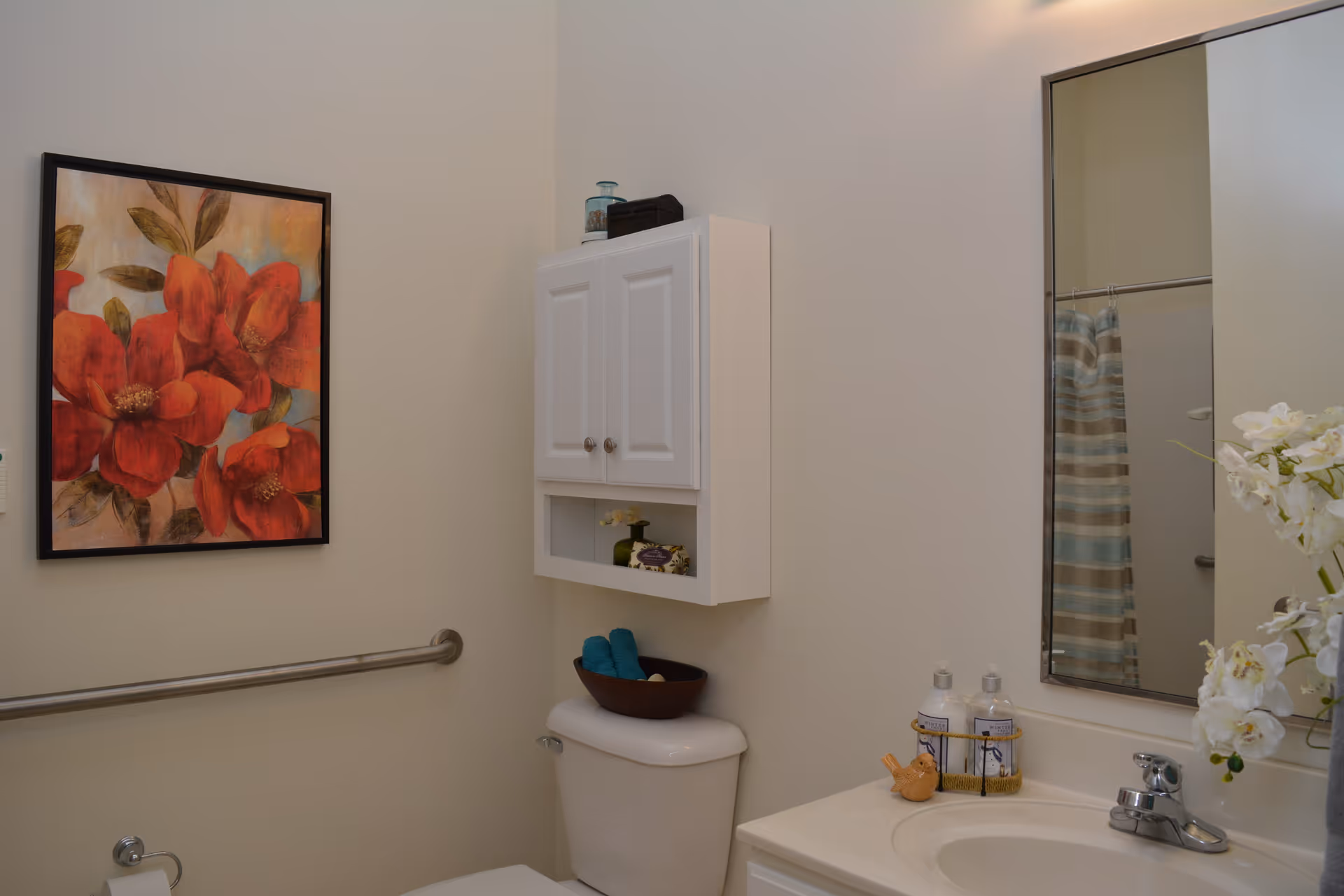 A bathroom with a white toilet, a white cabinet mounted above the toilet, a large mirror above a sink with a faucet, and a floral painting with red flowers on the wall. There is a grab bar on the wall next to the toilet and a striped shower curtain reflected in the mirror.