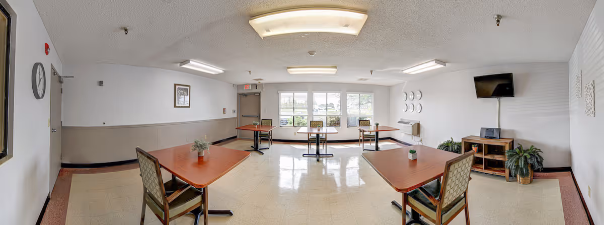 A bright and clean dining room with several wooden tables and chairs spaced apart. The room has large windows letting in natural light, a wall-mounted TV, potted plants, and decorative plates on the wall. The floor is tiled, and the walls are painted white with a beige lower half.