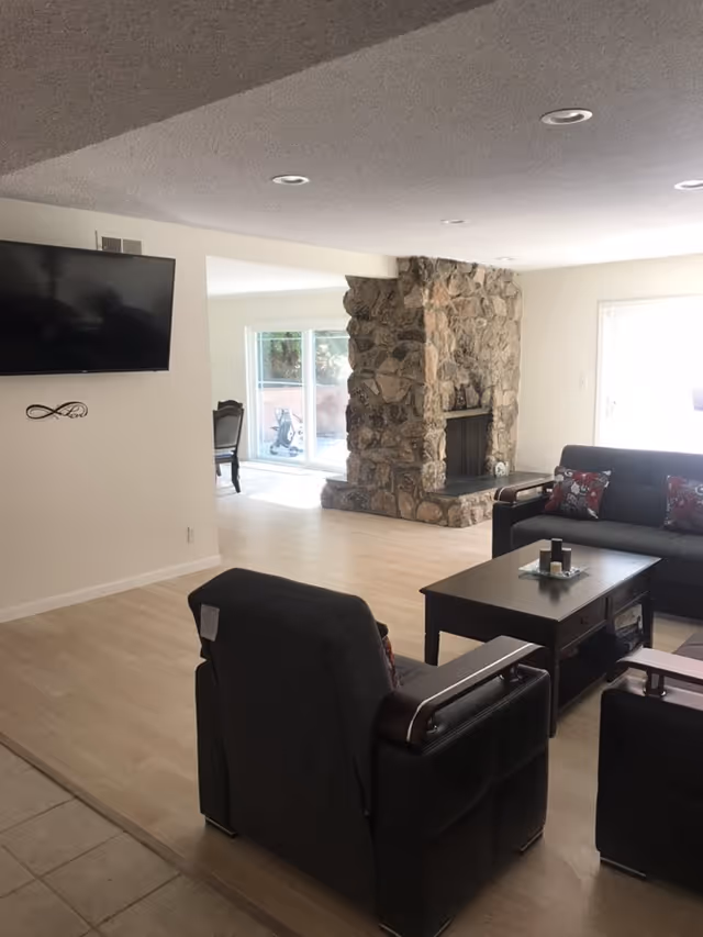 A living room with a stone fireplace, a dark gray sofa and armchairs with red patterned cushions, a dark wooden coffee table, and a wall-mounted flat-screen TV. The room has light wood flooring and white walls, with a dining area visible in the background through an open doorway.