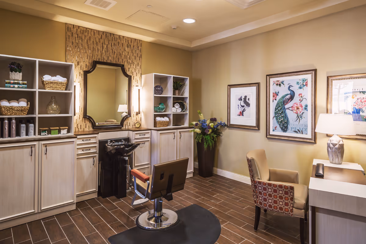 A well-lit salon room with a styling chair, wash station, mirror, cabinets, shelving, and framed artwork on the walls.