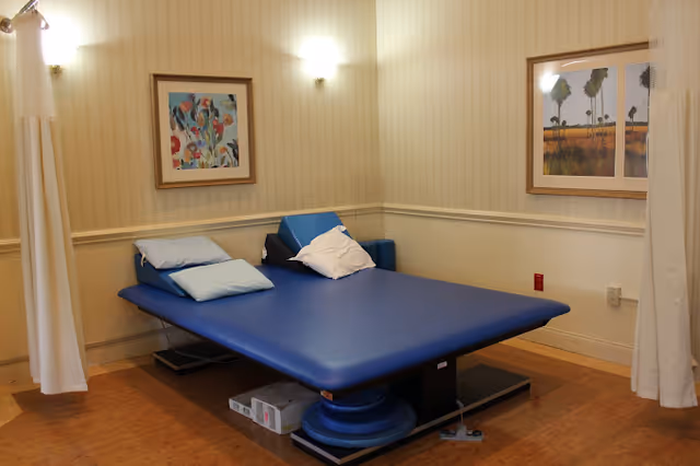 A therapy or treatment room with a large blue padded treatment table in the center, surrounded by light-colored walls with two framed paintings. The room has wooden flooring and is softly lit by wall-mounted lights. White curtains hang on either side of the table.