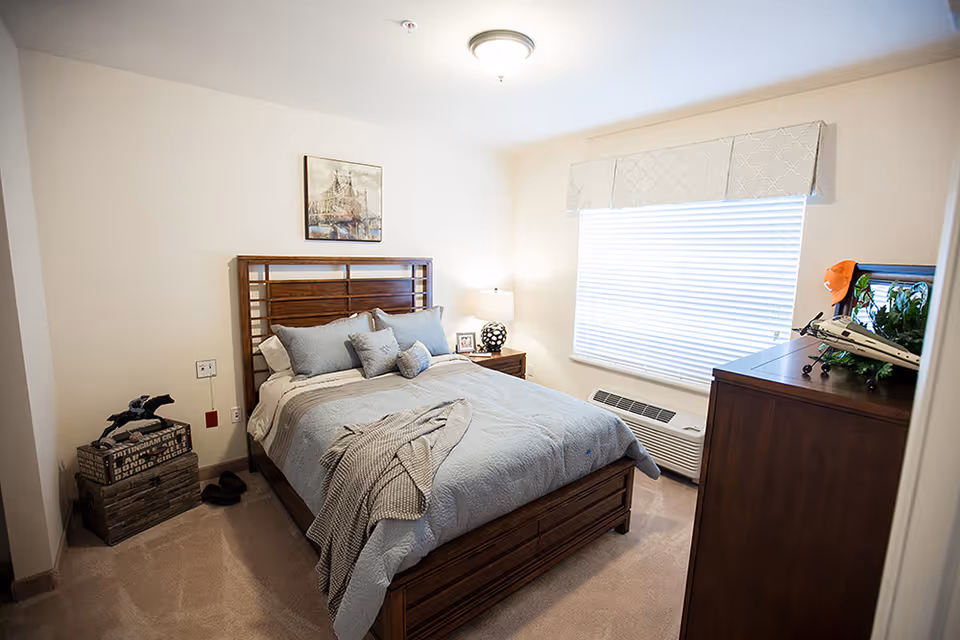 A cozy bedroom with a wooden bed frame and headboard, neatly made with light blue bedding and several pillows. A nightstand with a lamp and a framed photo is beside the bed. A window with blinds and a decorative valance lets in natural light. Across from the bed is a wooden dresser with a model airplane and a small plant on top. The room has beige carpet and neutral-colored walls with a framed picture hanging above the bed.